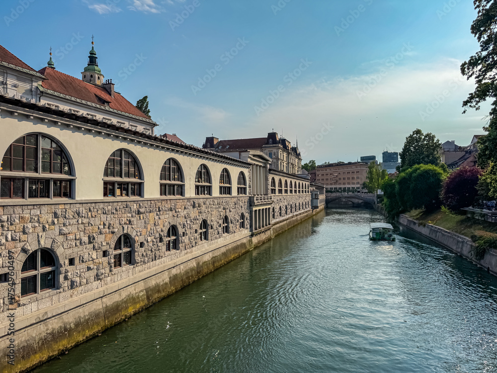 Fototapeta premium The stone arches of Plečnik's Central Market line the Ljubljanica river, where a tourist boat cruises past historic buildings under a blue sky on a perfect day for sightseeing in Ljubljana, Slovenia.