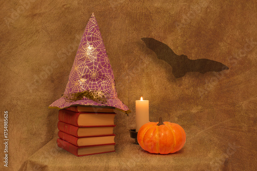 Halloween still life with several red books, a witch hat, a pumpkin, a white candle, and a bat, on a brown background