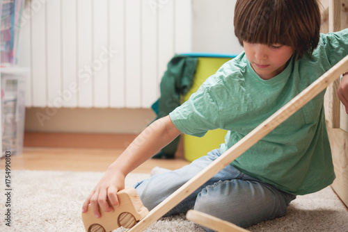 Young Caucasian boy explores motion by sliding a wooden van down a ramp while sitting on the floor. Natural play in a cozy home environment.