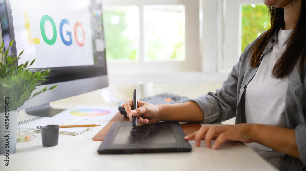 Fototapeta premium Graphic designer creating logo on digital tablet in bright modern workspace with computer and color palette
