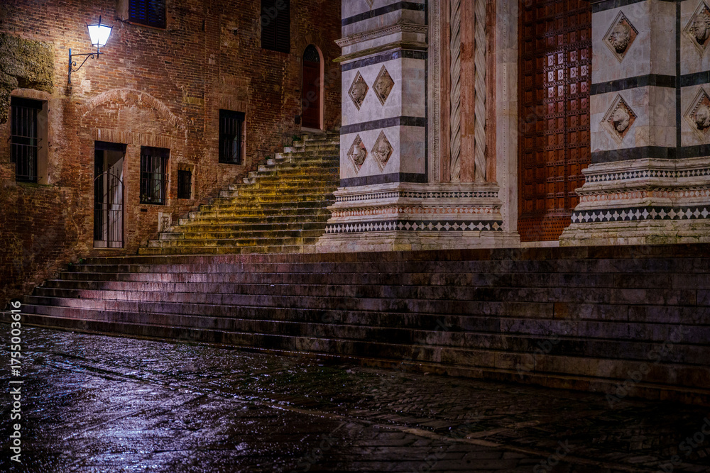 Naklejka premium Wet marble steps at Siena Cathedral entrance, ancient brick alley, night rain
