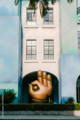 Modern architectural facade with a large golden hand sculpture making an “OK” gesture, framed by soft blue and white walls and greenery in the foreground