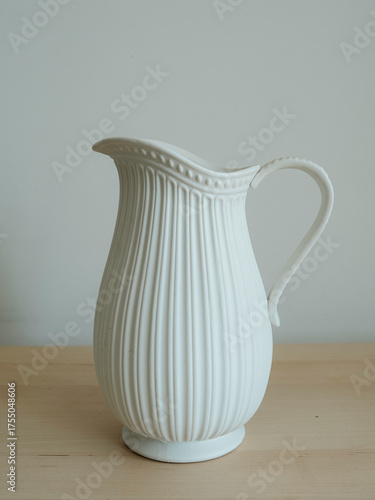 Minimalist photo of a white ceramic pitcher with elegant ribbed texture, placed on a wooden surface against a neutral background