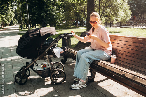 Young mom enjoys summer day in a park, sitting on a bench with a stroller, while drinking a refreshing cocktail and using her phone. Relaxed and multitasking in nature.