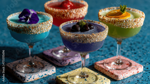 A row of colorful cocktails in martini glasses sit on a marble counter. The glasses are garnished with colorful sprinkles and fruit slices. The cocktails are arranged in a row