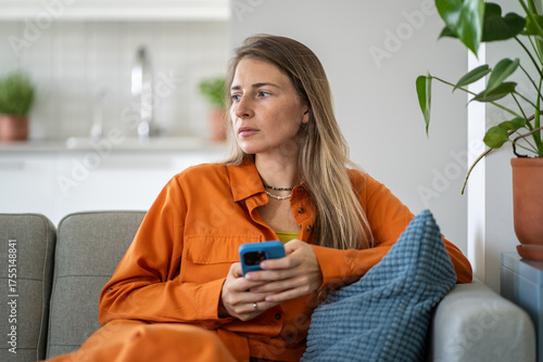 Obraz na plátne Pensive middle-aged woman frowning face in anxiety and stress holding phone seated alone on sofa