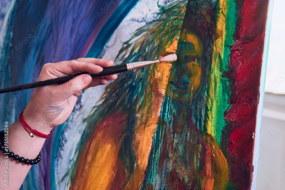 Fototapeta premium View of a woman's hands painting a picture. Next to the window, on a sunny day, multicolored paint, moving the brush, next to several brushes on her clothing, attitude, and beauty.