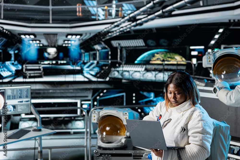 Fototapeta premium Female astronaut scientist operating spacecraft control interface using laptop. Woman researcher wearing cosmonaut gear in spaceship doing research, monitoring system diagnostics