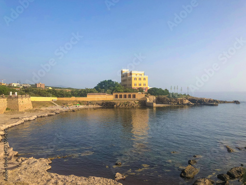Historisches gelbes Küstenschloss am Meer bei Alghero auf Sardinien, Italien, umgeben von Felsen und klarem Wasser unter blauem Himmel. Mediterrane Stimmung im Sommer.