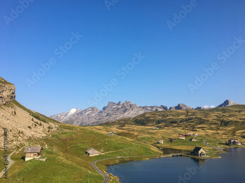 Idyllische Alpenlandschaft mit Bergsee, grünen Wiesen und verstreuten Hütten unter klarem blauem Himmel – friedliche Sommerstimmung in den Schweizer Alpen.