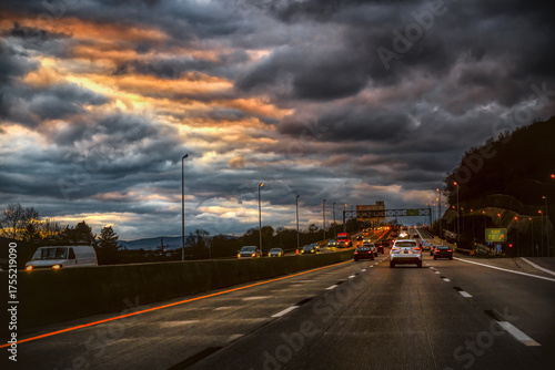 A dramatic sky with dark, fiery clouds hangs over a busy highway during the evening commute. The image captures the movement of vehicles on the interstate with bright orange and white lights.