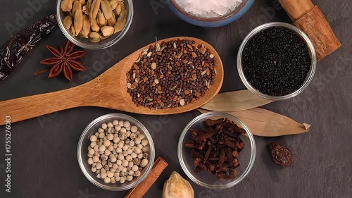 Overhead shot showcases colourful array of spices herbs on a wooden spoon pach phoron clove cardamom black seed salt pepper table spread of on dark black slate board