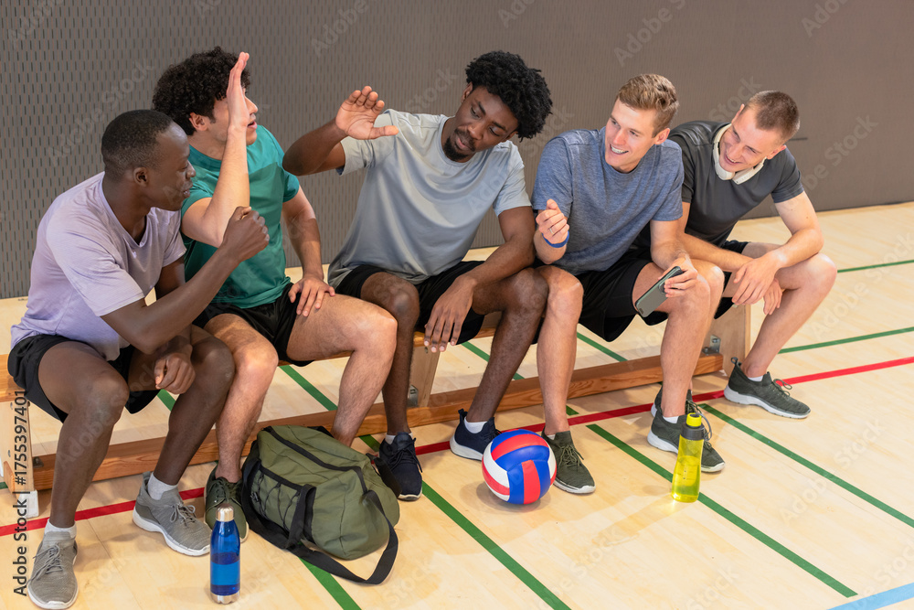 Obraz premium Diverse male volleyball players chatting and high-fiving on bench at gym with ball, duffel bag