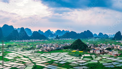 Fotografi Scenic view of a traditional village surrounded by reflective rice paddy fields at the base of karst mountains at sunset