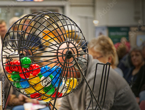 Bingo game cage at an event, colorful numbered lottery balls rotating inside as people watch, anticipating a draw