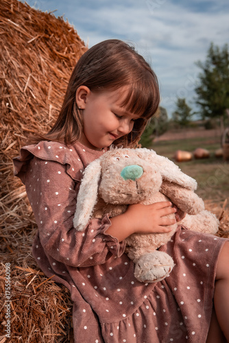 Happy little beautiful child girl hugs and plays with her favorite soft plush toy while sitting on a haystack in a park. Childhood. Autumn background.	