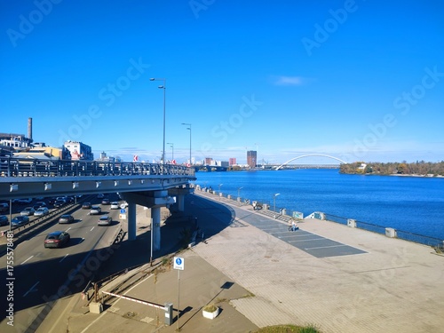 Sunny autumn day on the Dnipro River embankment in Kyiv. View of the river, bridges, and modern urban development on the left bank. Bright blue sky and water.
