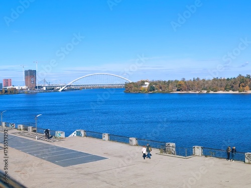 Sunny autumn day on the Dnipro River embankment in Kyiv. View of the river, bridges, and modern urban development on the left bank. Bright blue sky and water.