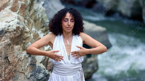 A woman in a flowing white outfit performs an expressive dance by a tranquil riverside, surrounded by rocky landscapes that enhance her fluid movements.
