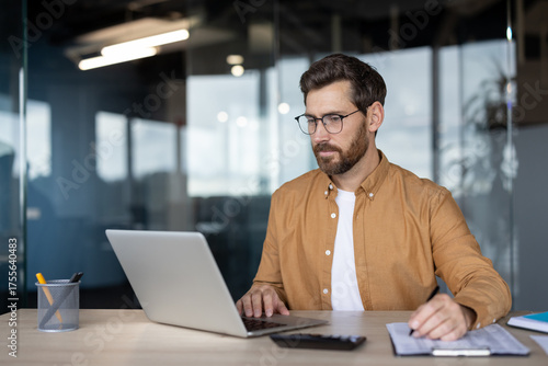 Businessman in glasses and a brown shirt is actively working at his office desk, typing on a laptop and writing notes, demonstrating focus on work, productivity, and professional multitasking
