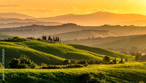 Lush green hills cascade down to a valley floor under a glowing golden sunset sky with rolling mountains