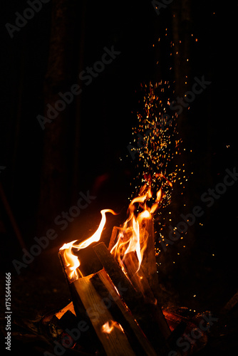 Dramatic vertical shot of a bright campfire burning stacked wood in the darkness. A stream of glowing embers shoots upward, creating a vibrant, dynamic, and moody effect.