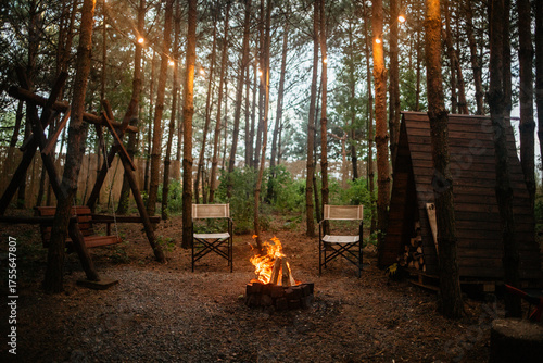 Foto A warm, inviting campsite scene at dusk with a blazing campfire, two empty chairs, a rustic wooden swing, and a firewood shed, all illuminated by string lights hanging in the pine trees