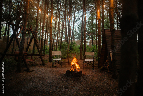 Cozy forest campsite at twilight featuring a brightly burning campfire and two canvas folding chairs. String lights hang above, creating a warm, inviting atmosphere among the pine trees.
