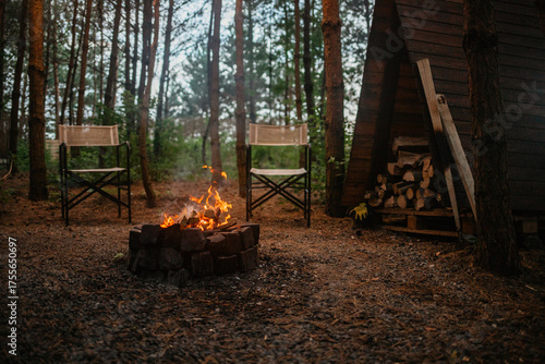 A wide, cozy glamping scene at dusk featuring a blazing stone fire pit and two empty canvas chairs, with a rustic A-frame wood storage shed in a quiet pine forest setting.