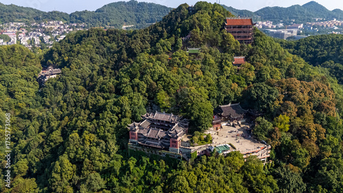 Aerial view of a historic Chinese temple complex with traditional architecture, beautifully situated amidst the dense green forest of a mountain in Chongqing, with the city in the distance