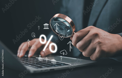 Businessman analyzing financial data and interest rates through a magnifying glass while using a laptop. Concept of banking, investment research, economy growth, and digital finance strategy.