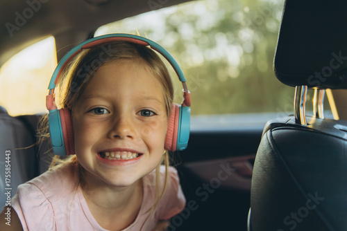 Smiling girl wearing colorful headphones sitting in the back seat of a car during road trip. Happy child enjoying music while traveling.