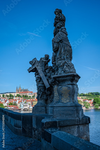 Sculpture of Socha svaté Anny on Charles Bridge over the Vltava River in Prague Old Town, Czech Republic, with Prague Castle visible in the distance under a clear blue sky.
