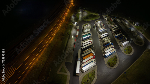 Aerial view of illuminated truck stop at night with parked vehicles along highway, providing respite.