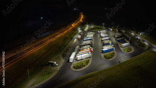 Aerial night view of trucks parked in a rest area next to a brightly lit highway, dark background
