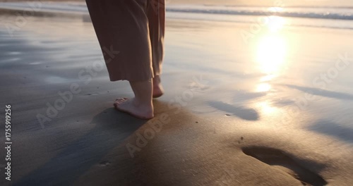 Slow motion close-up of a woman's bare feet walking on wet sand. The golden sun reflects on the shoreline as she leaves footprints.