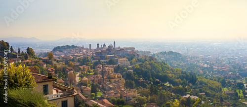 A panoramic view of Bergamo with numerous buildings and a hill in the background. The sky is overcast and the sun is shining.