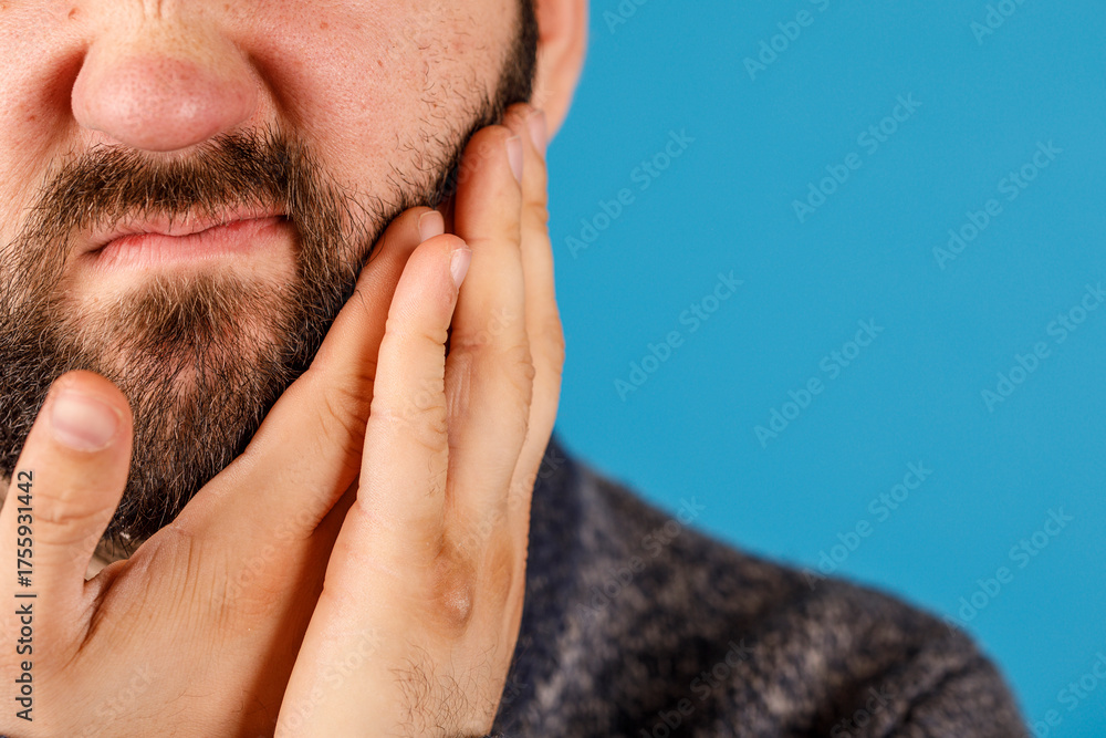 Obraz premium Close-up of a man holding his face, possibly in pain, with a blue background. Perfect for health, wellness, or discomfort themes.