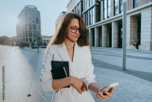 Photography A woman in glasses glances at her phone, surrounded by towering modern buildings in a lively city