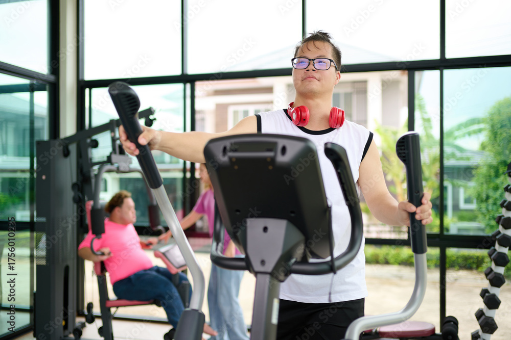 Fototapeta premium A man with down syndrome exercising on an elliptical machine in a modern gym setting with others working out in the background