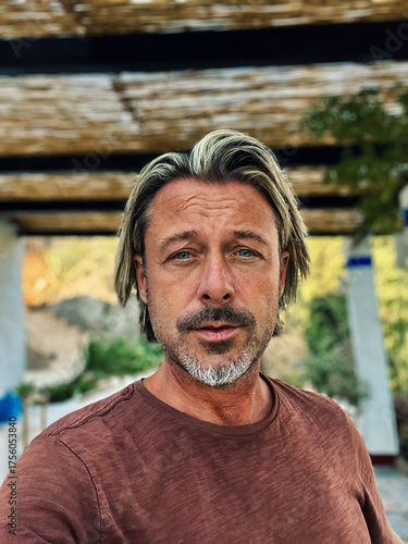 Man with half long blond hair and grey beard wearing brown t-shirt outside under a veranda in spanish countryside during summer.