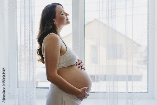 A Pregnant woman stands by a window, cradling her round belly. This moment captures the beauty of pregnancy. Free Download A Pregnant woman stands by a window, cradling her round belly. This moment captures the beauty of pregnancy. - GFXLoad