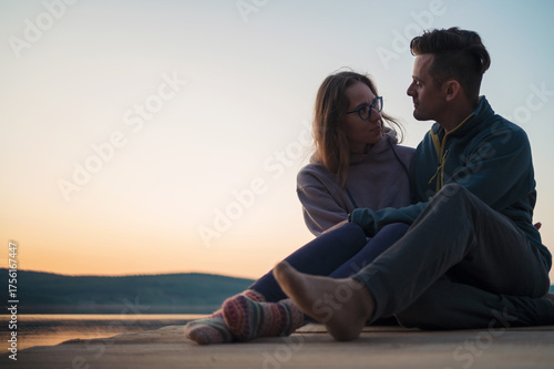 Loving couple sitting close together on a wooden pier by the calm lake at sunset, enjoying peaceful evening moments and connection.