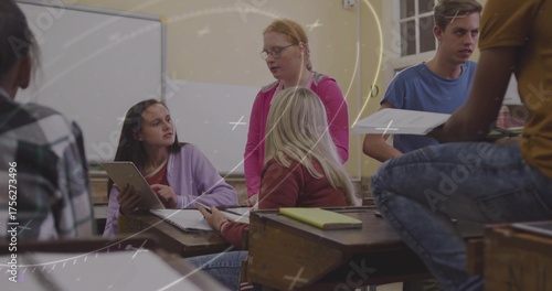 Collaborating students pointing at tablet screen in classroom, passing papers at wooden desks