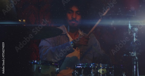 Playing drummer wearing light denim shirt over white T-shirt in dimly lit stage, with drum kit
