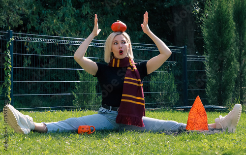 A beautiful blonde girl is sitting on the grass, holding a Halloween pumpkin on her head