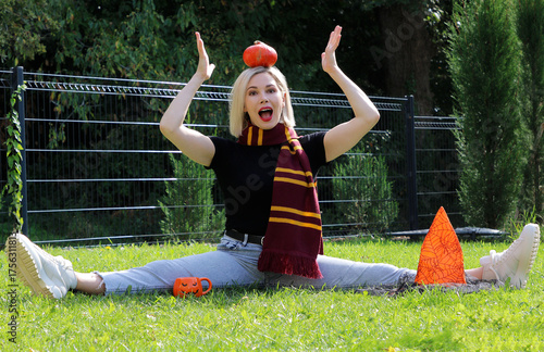 A beautiful blonde girl is sitting on the grass, holding a Halloween pumpkin on her head