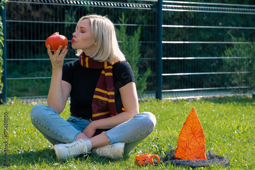 A beautiful girl in an autumn scarf is sitting on the grass and kissing a Halloween pumpkin