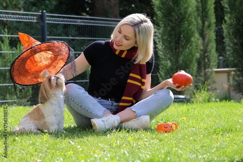 A beautiful girl is sitting on the green grass, putting a Halloween hat on a dog.