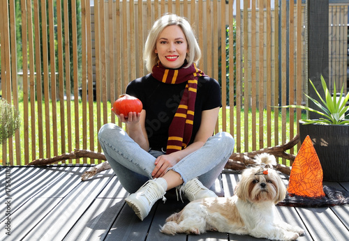 A beautiful blonde girl is sitting on the terrace with a dog, holding a Halloween pumpkin in her hands